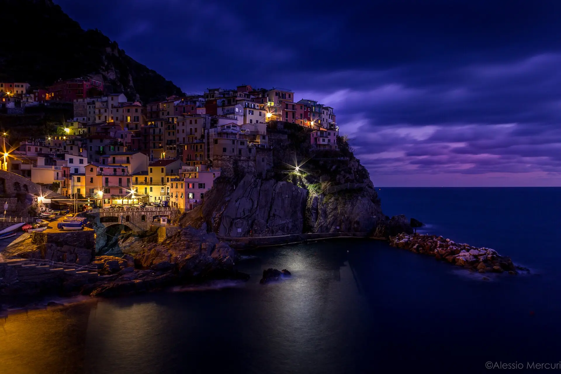 Manarola at twilight from the Nessun Dorma viewpoint — village lights along the cliffs reflected in the harbour at blue hour.