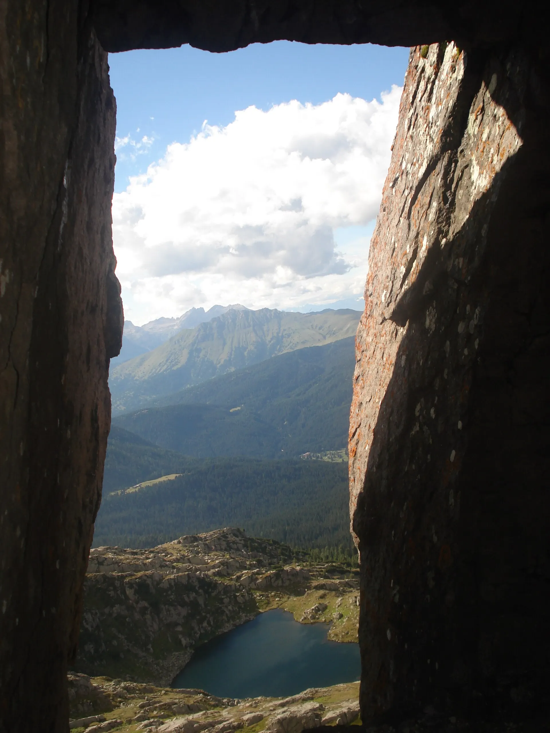 Looking down through a rock notch on the Monte Cavallazza ridge to a small alpine lake below, with the Lagorai range stretching to the horizon.