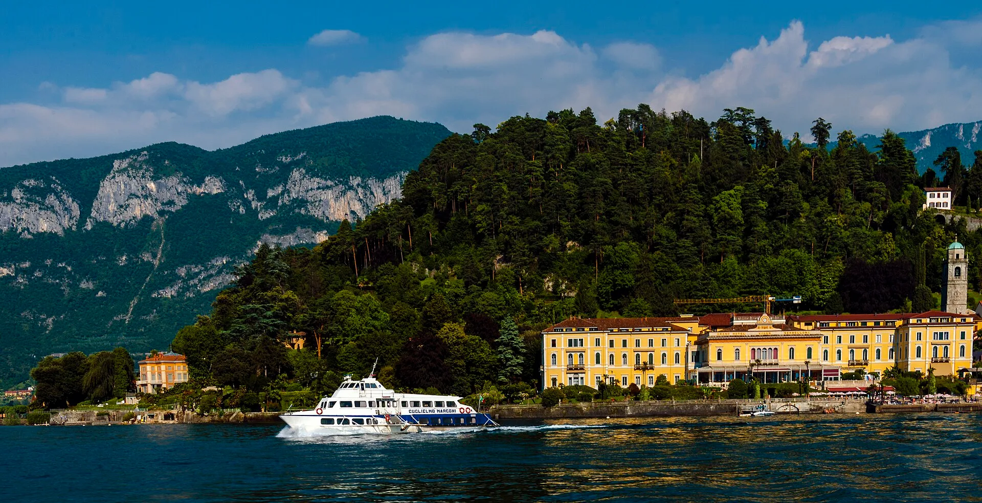 Bellagio waterfront on Lake Como, seen from a passing ferry — yellow palazzi above the lake, wooded headland behind.