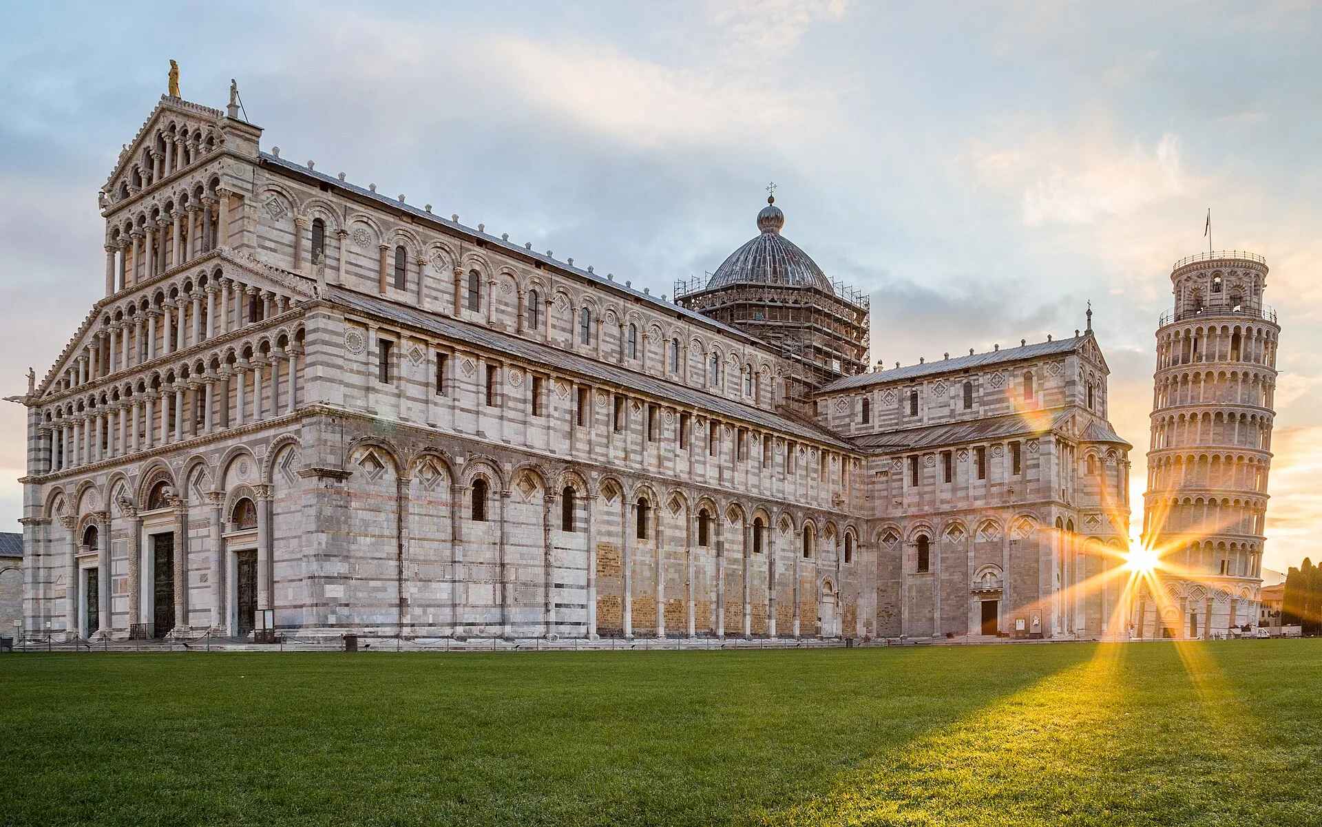 Pisa Cathedral and the Leaning Tower at sunset, with a sunburst between them on the Piazza dei Miracoli.